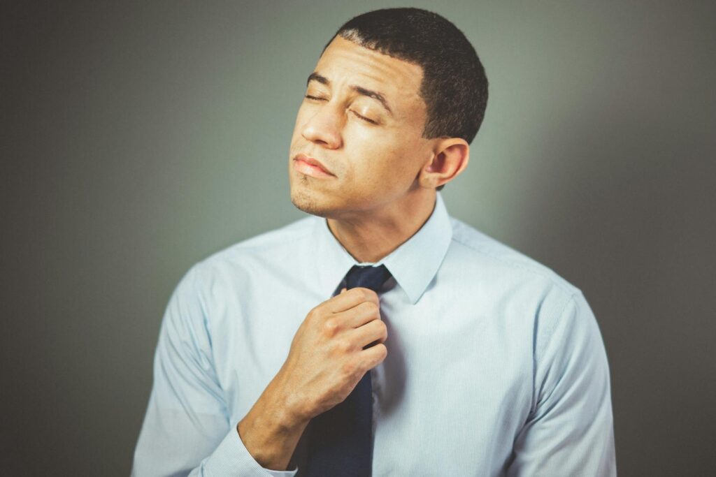 A man indoors adjusting his necktie with a thoughtful expression, wearing formal attire.
