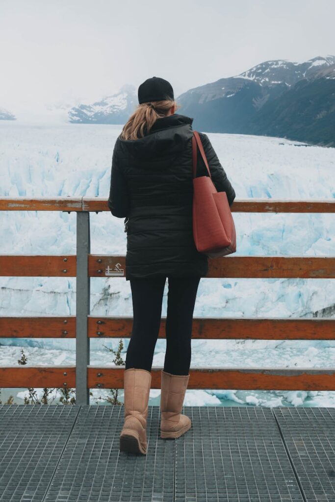 Woman in warm attire standing on a platform, overlooking a stunning glacier.