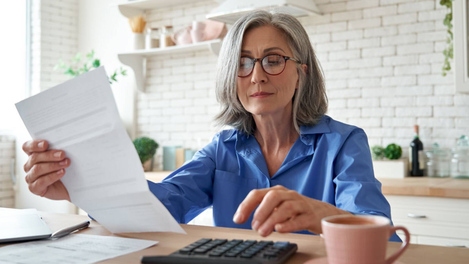 A woman is seen sitting at a kitchen table with a calculator looking at the Social Security benefits she will receive in retirement. -lead